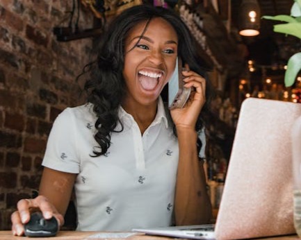 A young woman sitting at a laptop smiles and laughs on the phone